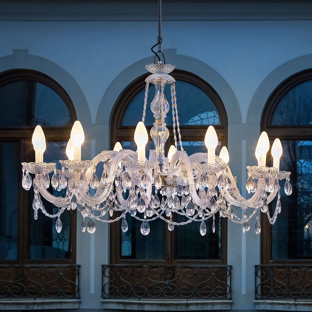 An Outdoor Cut Crystal Chandelier gracefully hangs, casting light on an old stone building with arched windows. Its reflection shimmers in an empty pool, creating a serene and luxurious ambiance.
