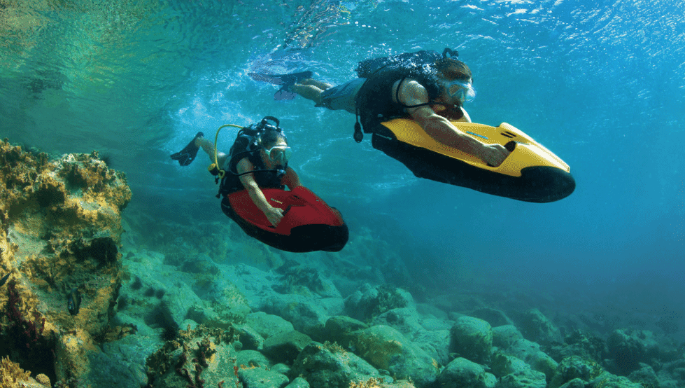 Yacht toys, two people underwater with red and yellow seabob, coral reef underneath