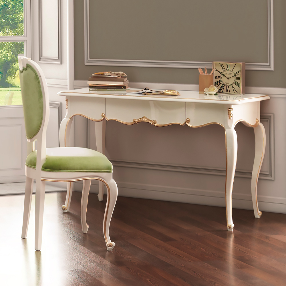 A vintage-style writing desk with ornate legs and light wood finish is situated against a wall with classic paneling. On the desk are a clock, books, and stationery. A matching chair with light green upholstery stands in front of the desk on a polished hardwood floor.