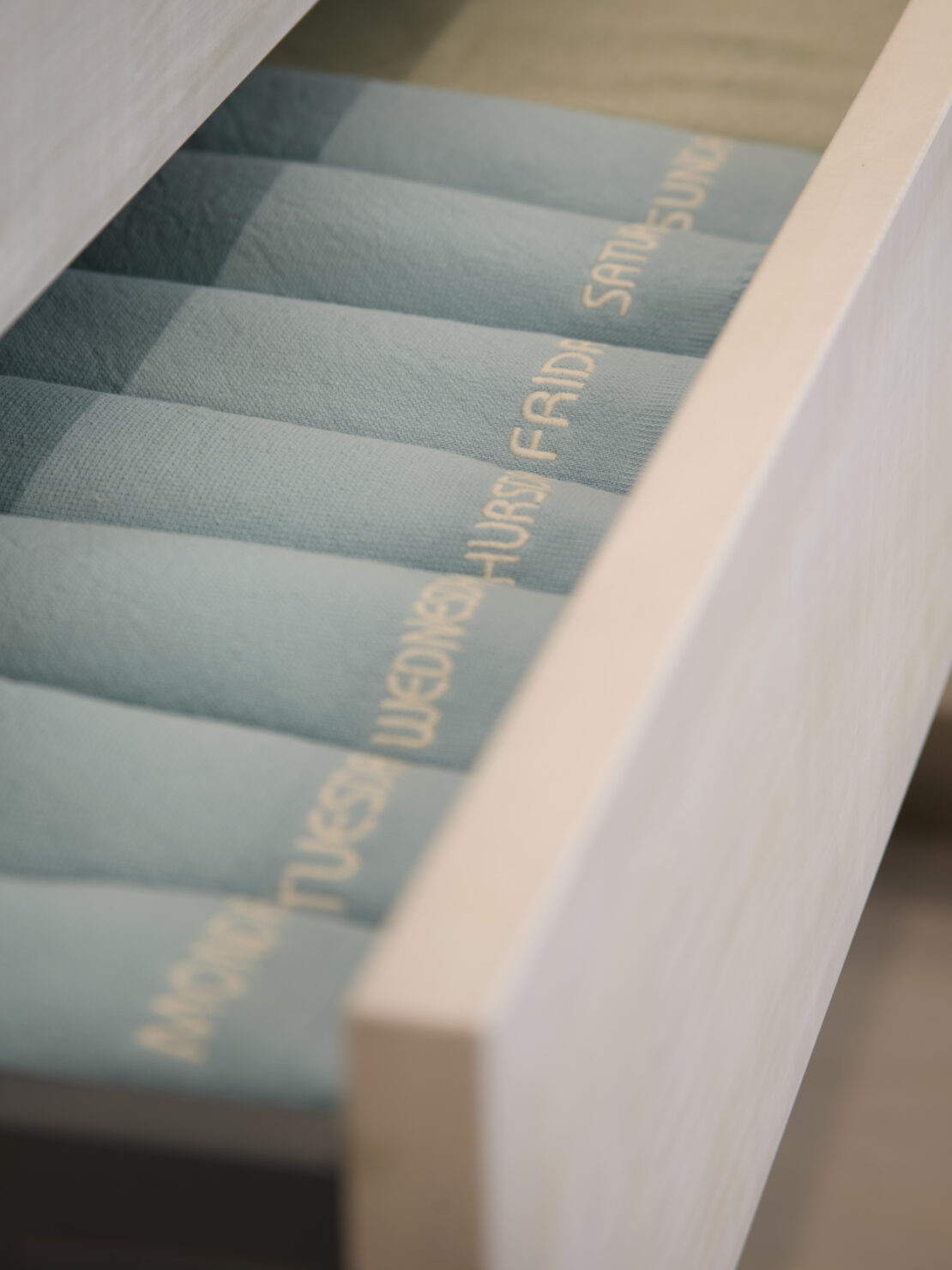 A partially open drawer in the stylish Battersea apartment reveals neatly rolled light blue and green socks, each embroidered with the days of the week: Monday, Tuesday, Wednesday, Thursday, Friday, Saturday, and Sunday, all arranged in perfect order.