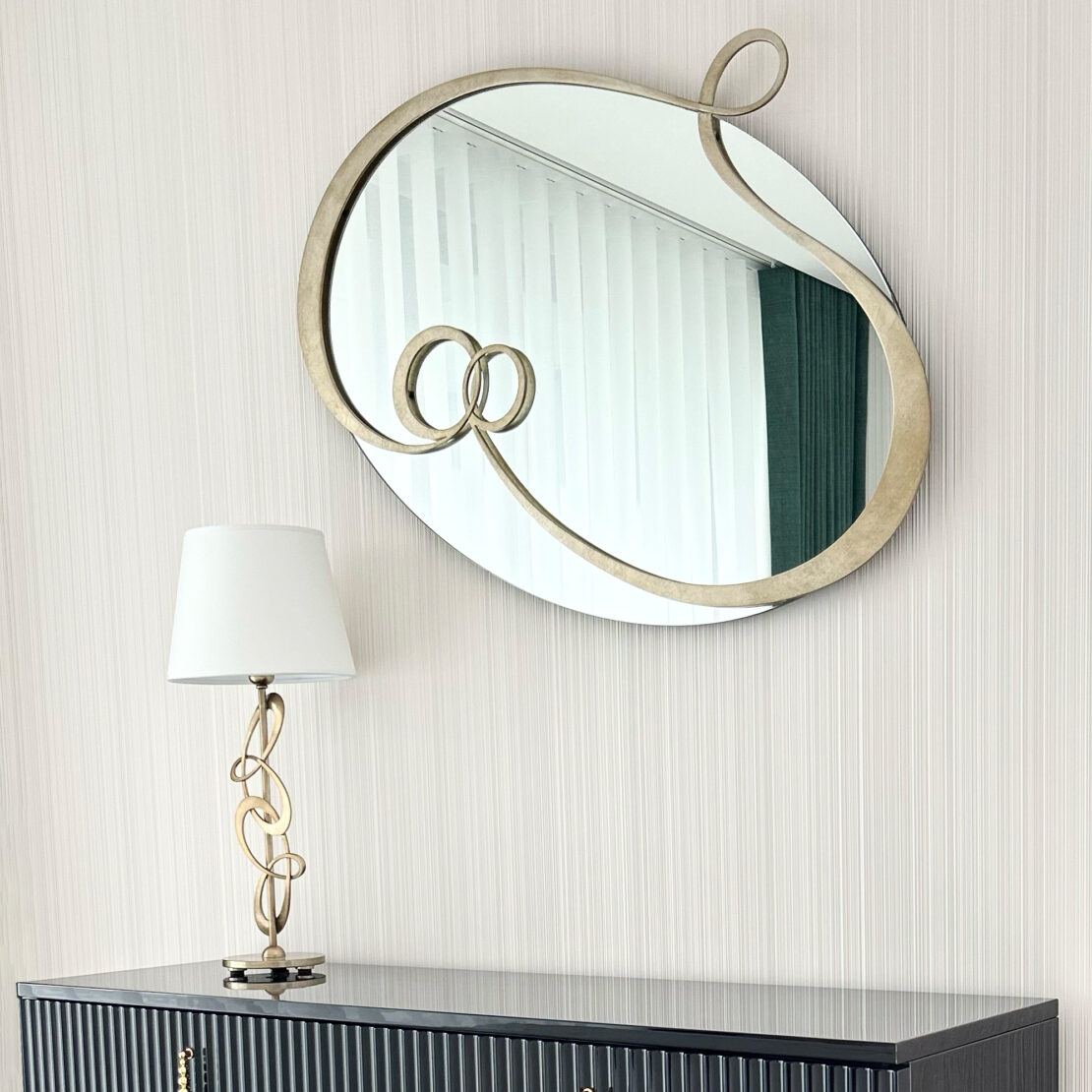 Contemporary Twisted gold mirror. A round mirror with a gold, looped frame hangs on a light-colored wall above a dark, ribbed sideboard. The sideboard features brass handles and is topped with the Contemporary Twisted Table Lamp, which has a matching gold looped design and is capped with a white shade.