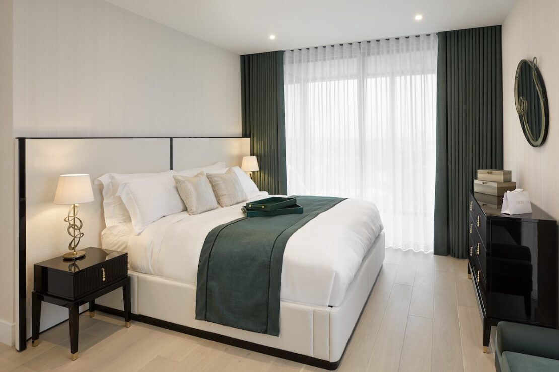 A modern bedroom design with a neatly made bed featuring white linens and green accents, including a throw and pillows. The room has matching green curtains, a bedside table with a lamp, a black dresser with decor items, and a round mirror on the wall. The floor is light wood.