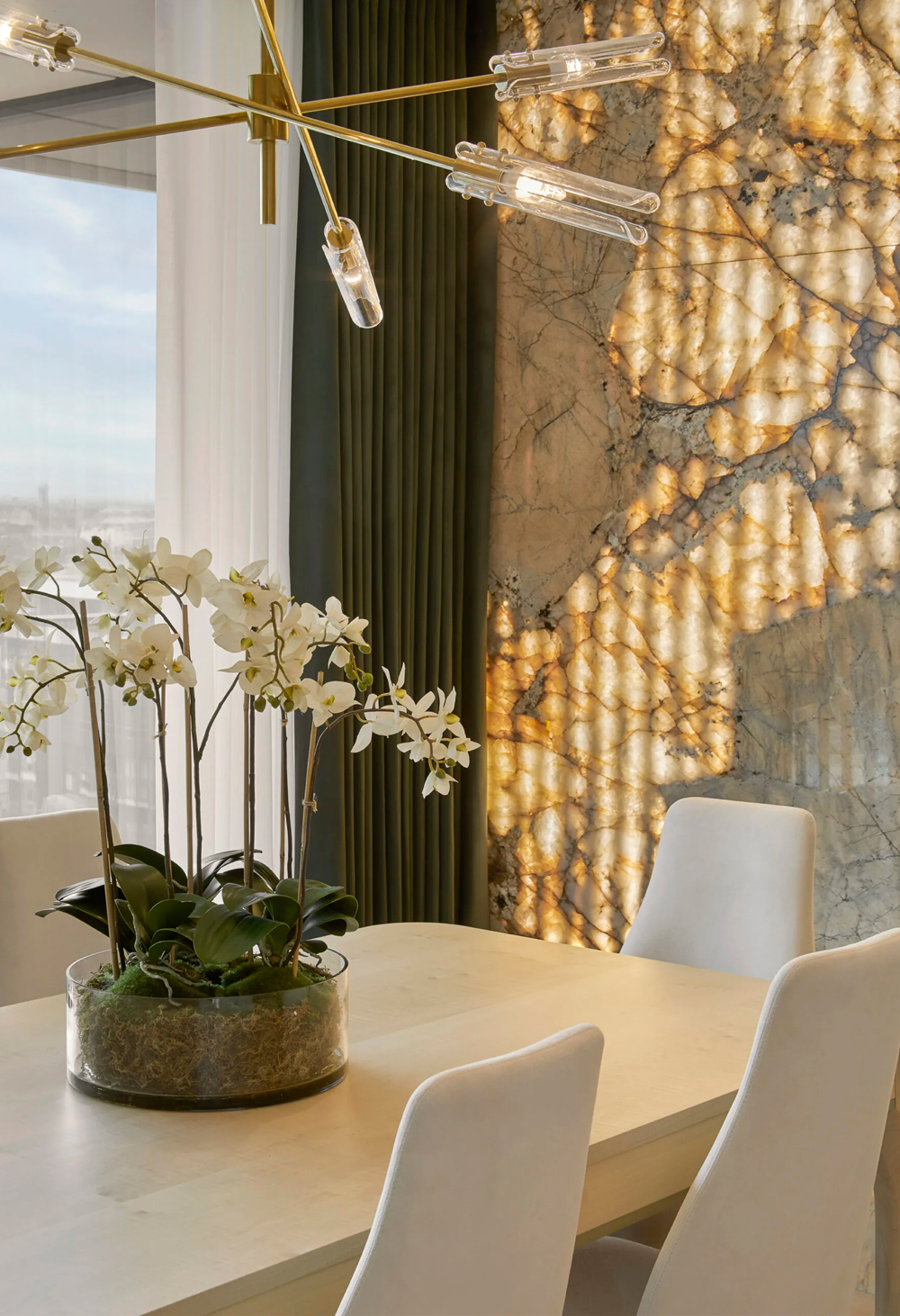A modern dining area in this Apartment Battersea features a light wood table with white chairs. A glass pot of white orchids serves as the centerpiece. The wall showcases a striking backlit marble design, while a contemporary chandelier with exposed bulbs hangs above the table.